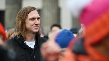 Ein Mann mit schulterlangen Haaren steht in einer nur unscharf erkennbaren Menschenmenge.