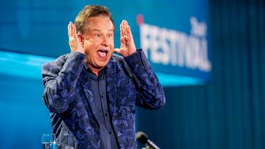 Kabarettist steht auf der linken Bildseite. Er hebt beide Hände neben dem Kopf. Sein Mund ist offen. Er trägt einen blauen Anzug mit Muster und darunter ein dunkelblaues Hemd. Die Bühne ist blau beleuchtet.