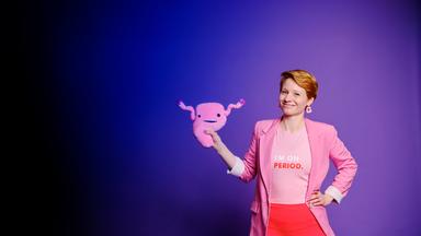 Steffi Platt steht vor einer lilablauen Wand. Sie hat eine orangefarbene Leggings und einen rosa Blazer an. Außerdem trägt sie ein T-Shirt mit der Aufschrift: "I‘m on period.". In der rechten Hand hält sie eine Gebärmutter aus Plüsch. Ihren linken Arm hat sie in die Hüfte gestemmt. Sie lächelt in die Kamera.