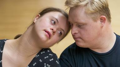 Das Bild zeigt zwei junge Menschen, die in einem persönlichen Moment des Tanzes zu sehen sind. Die Frau hat lange, dunkle Haare und trägt ein schwarzes Oberteil mit einem Muster aus kleinen weißen Insekten. Sie lehnt sich leicht an den Mann, der kurzes, blondes Haar hat und ein schwarzes T-Shirt trägt. Beide wirken nachdenklich, ihre Gesichter sind nah beieinander, und sie schauen sich intensiv an. Der Hintergrund ist unscharf, mit einem hellen Holzboden, der den Eindruck eines Tanzraums vermittelt. Das Bild strahlt eine emotionale Verbindung und Intimität zwischen den beiden Protagonisten aus.