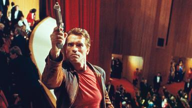 Actionstar Jack Slater (Arnold Schwarzenegger) steht mit gezückter Pistole auf den Bänken des ersten Rangs und visiert einen imaginären Feind an. Hinter ihm das gut gekleidete Premierenpublikum im Kinosaal.