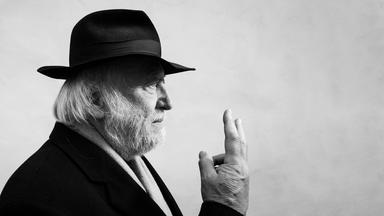 Schwarzweißes Profilfoto eines älteren Mannes mit grauen Haaren und Bart, der einen schwarzen Hut trägt und die rechte Hand mit zu einem V gespreizten Zeige- und Mittelfinger vor sich hält.