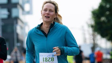 Juliane (Anna Schudt) joggt in einer blauen Laufjacke, mit einer vierstelligen Startnummer auf dem Bauch, auf einer Straße entlang.