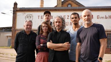 Sechs Personen stehen vor einem alten, industriell anmutenden Gebäude. Der Mann links trägt ein schwarzes Hemd und hat graues Haar. Neben ihm steht eine Frau mit langen, rötlichen Haaren, die ein gemustertes Oberteil und eine rotbraune Hose trägt. Daneben ist ein Mann mit mittellangem, grauem, lockigem Haar und schwarzer Kleidung, der die Arme verschränkt hat zu sehen. In der hinteren Reihe stehen von links nach rechts ein Mann mit schwarzer Kappe und schwarzem Hemd, ein jüngerer Mann mit kurzem, dunklem Haar in einem blauen T-Shirt und ein älterer Mann mit kahlrasiertem Kopf und einem dunkelblauen T-Shirt