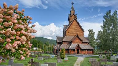Mittelalterliche norwegische Stabkirche aus Holz mit steilen Dächern, umgeben von grüner Natur und Bergen im Hintergrund.