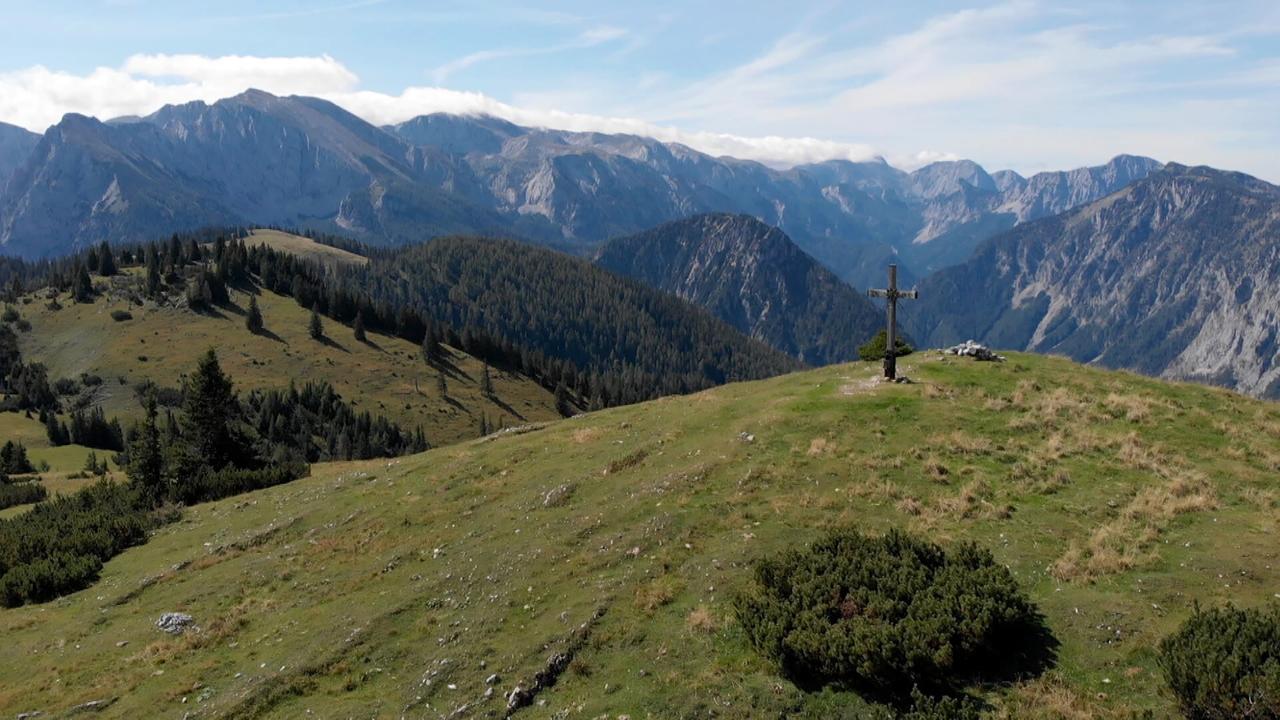 Das Bild zeigt eine weite, alpine Landschaft im Mariazellerland. Im Vordergrund erstreckt sich eine grüne Wiese mit vereinzelten Sträuchern und Steinen. In der Mitte der Bildfläche steht ein hölzerner Kreuz, das als religiöses Symbol platziert ist.   Im Hintergrund erhebt sich eine beeindruckende Gebirgskette mit steilen, felsigen Hängen und bewaldeten Bereichen. Die Berge sind von einer blauen Himmelspanne umrahmt, die teilweise mit hellem, fluffigem Wolkenbewuchs versehen ist.   Insgesamt strahlt die Szene eine ruhige und naturnahe Atmosphäre aus, die die alpine Prägung der Region unterstreicht. Es gibt keine menschlichen Figuren oder Gebäude im Bild.