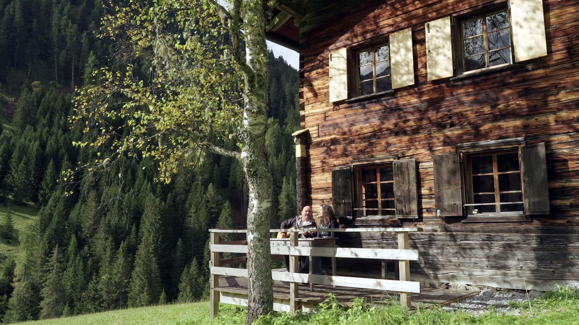 Das Bild zeigt eine Almhütte in einer bergigen Landschaft. Die Hütte ist aus Holz gebaut und hat große Fenster mit dunkelbraunen Fensterläden, die teilweise geöffnet sind. Vor der Hütte befindet sich eine Holzveranda, auf der zwei Personen sitzen. Die eine Person hält eine Gitarre. Im Hintergrund erstrecken sich dichte, grüne Nadelwälder, die die Berge umgeben. Der Himmel ist teilweise bewölkt, und die Sonne scheint durch die Bäume, was Licht- und Schattenspiele auf die Holzfassade der Hütte erzeugt. Es scheint eine ruhige, ländliche Atmosphäre zu herrschen.