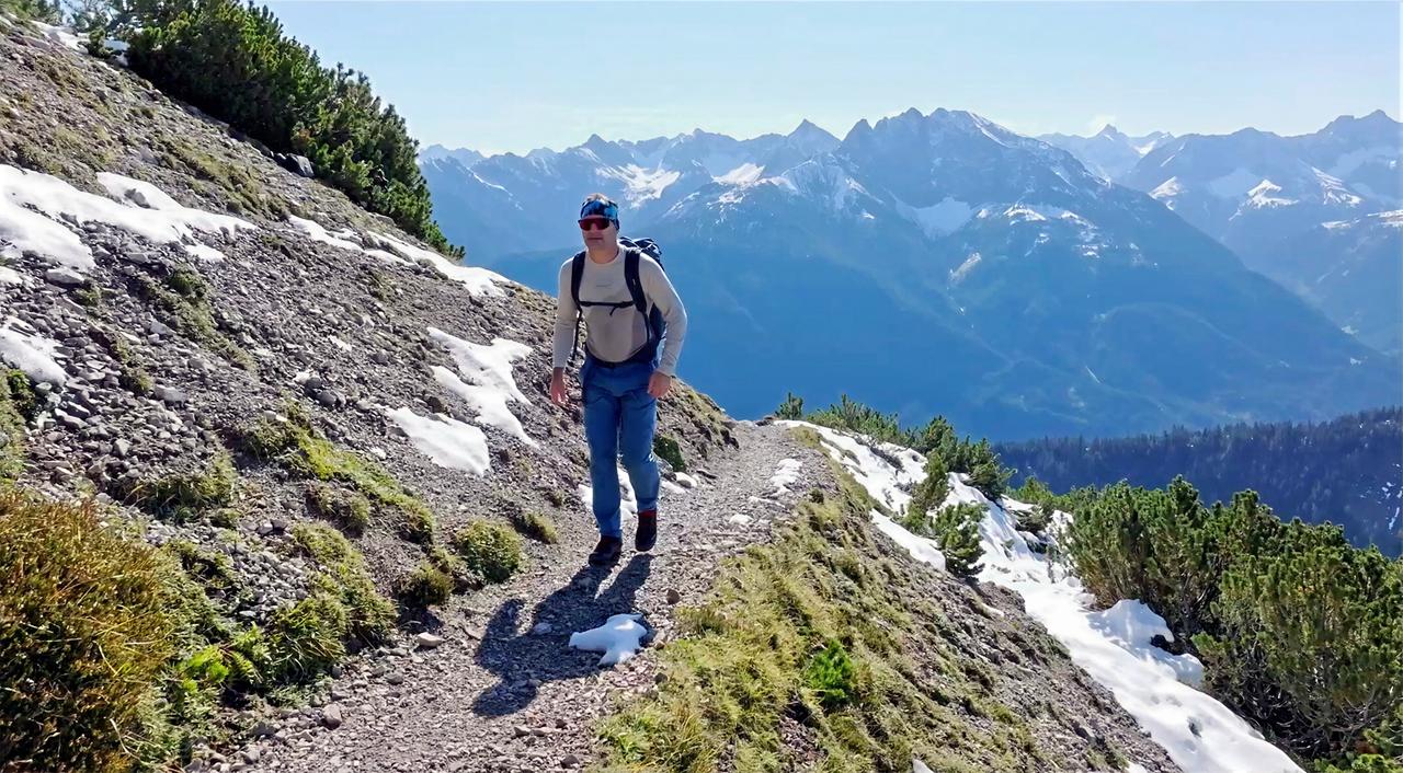 Das Bild zeigt eine bergige Landschaft im Tiroler Lechtal. Im Vordergrund ist ein Wanderweg, der sich entlang eines Hang mit steinigem Untergrund schlängelt. Reste von Schnee sind auf dem Weg und an den Seiten sichtbar, während dichter, grüner Bewuchs, wie kleinwüchsige Bäume und Sträucher, den Hang ziert.  Auf dem Weg befindet sich ein männlicher Wanderer. Er trägt eine helle, langärmlige Oberbekleidung, eine leichte Jacke, eine Sporthose und hat einen Rucksack auf dem Rücken. Auf seinem Kopf trägt er eine Sonnenbrille und eine Kappe. Der Wanderer bewegt sich in Richtung einer Hütte, die im Hintergrund weiter oben im Bild zu vermuten ist.  Im Hintergrund des Bildes sind hohe, majestätische Berge zu sehen, die zum Teil mit Schnee bedeckt sind, und der Himmel ist blau mit wenigen Wolken. Die Landschaft strahlt Ruhe und natürliche Schönheit aus.