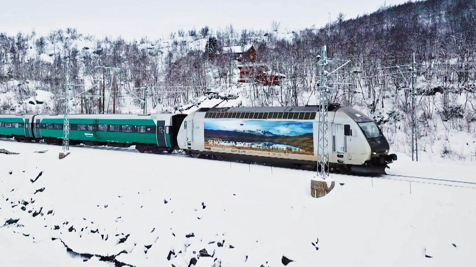 Ein Zug mit grünen Waggons und einem weißen Triebwagen, auf dem ein Bild von einer norwegischen Berglandschaft abgebildet ist, fährt durch eine Schneebedeckte Landschaft.