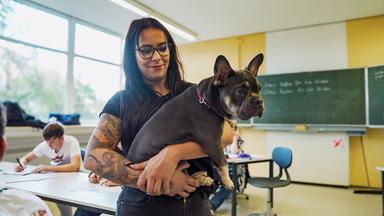 Eine Frau mit einem Hund auf dem Arm in einem Klassenzimmer.