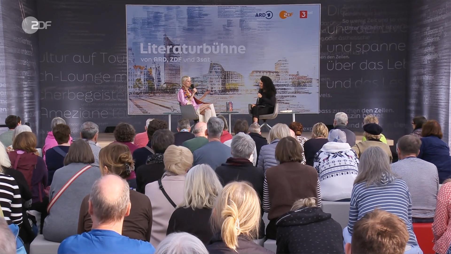 Zwei Frauen sitzen auf einer Bühne vor Publikum und unterhalten sich. Im Hintergrund ist ein Bild eingeblendet, auf dem Lesebühne steht.