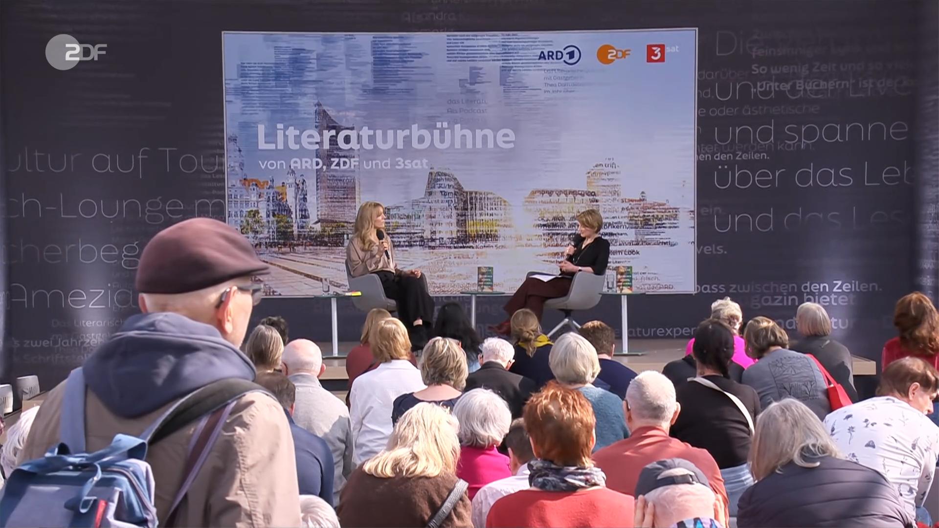 Zwei Frauen sitzen auf einer Bühne vor Publikum und unterhalten sich. Im Hintergrund ist ein Bild eingeblendet, auf dem Lesebühne steht.