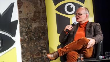 Ein Mann mittleren Alters sitzt auf einem Podium mit dem Logo der Leipziger Buchmesse im Hitergrund.
