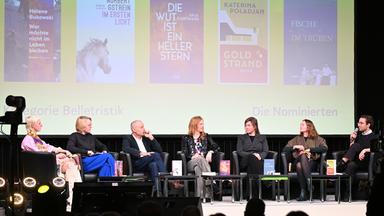 Auf einer Bühne sitzen die sechs nominierten für den Leipziger Buchpreis. Im Hintergrund werden ihre Bücher eingeblendet.