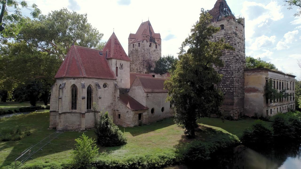 Das Bild zeigt eine historische Schlossanlage, umgeben von Grünflächen und Bäumen. Auf der linken Seite ist ein Gebäude mit einem hohen, markanten Schornstein und mehreren spitzen Dächern sichtbar. Die Dächer sind rot und aus Ziegeln, während die Wände des Schlosses aus grauem Stein bestehen, der an einigen Stellen verwittert ist. Es gibt große Fenster, einige davon mit Rundbögen.  Rechts im Bild ist ein weiteres, flacheres Gebäude zu sehen, das auch aus Stein besteht und teilweise von Pflanzen überwuchert ist. Der Vordergrund des Bildes ist von grünem Gras und Sträuchern geprägt, und ein schmaler Wasserlauf verläuft am unteren Bildrand. Der Himmel ist teilweise bewölkt, mit etwas Blau zwischen den Wolken.   Das Schloss hat historische Bedeutung und ist Teil der Erbe-Österreich-Reihe, die sich mit Wasserwegen und deren Geschichte befasst.