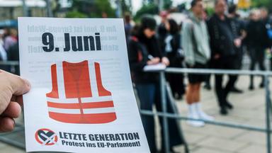 Eine Hand hält einen Flyer der Letzten Generation für die Wahl zum EU-Parlament am 9. Juni. Im Hintergrund sind viele Menschen verschwommen zu erkennen.