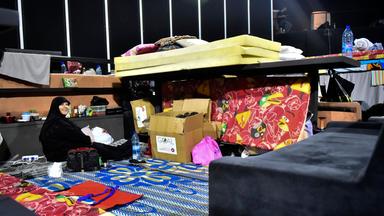 Eine Frau in schwarzer Kleidung und mit schwarzem Kopftuch sitzt auf dem Boden auf bunten Teppichen zwischen Kartons und Matratzen in einem dunklen Raum.