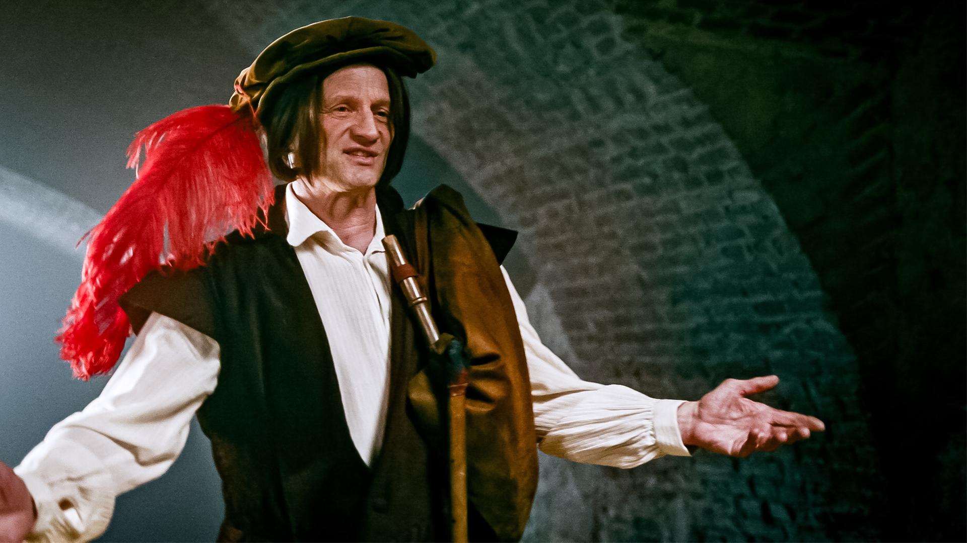 Das Bild zeigt einen Mann in historischer Kleidung, der auf einer Bühne steht und eine Art Performance oder Ansprache hält. Er trägt ein braunes Gewand mit einem weißen Hemd und einen Hut, der mit einer roten Feder verziert ist. Der Mann hat mittellange, leicht lockige Haare und schaut fröhlich, während er mit beiden Händen gestikuliert.   Im Hintergrund sind gewölbte Wände aus Ziegelstein sichtbar, die eine dunkle, aber stimmungsvolle Atmosphäre schaffen. Es scheint, dass die Szene in einem Innenraum spielt, möglicherweise in einem alten Gebäude oder unterirdischen Raum. Die Beleuchtung ist gedämpft und fokussiert auf die Person, wodurch ein Gefühl von Dramatik und Intensität erzeugt wird.   Das Bild stammt aus einem Kontext, der die Geschichte des „lieben Augustin“ erzählt, einer Figur aus Wien im Jahr 1679, die während der Pestzeit lebt.