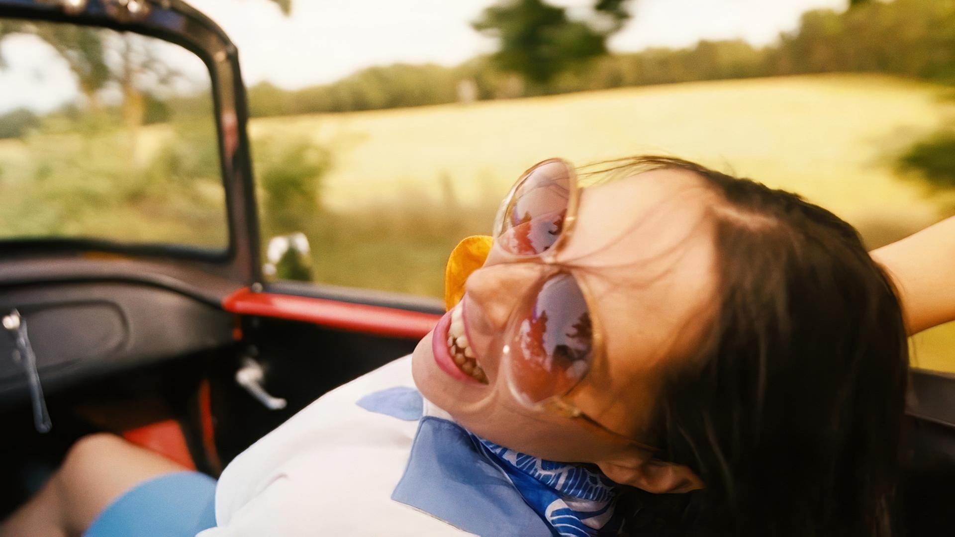 Eine Frau mit Sonnenbrille lehnt sich in einem offenen Cabrio zurück.