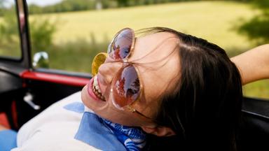 Eine Frau mit Sonnenbrille lehnt sich in einem offenen Cabrio zurück.