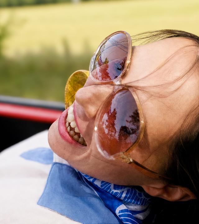 Eine Frau mit Sonnenbrille lehnt sich in einem offenen Cabrio zurück.