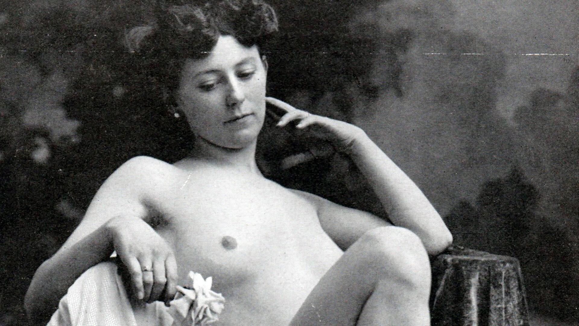 Das Bild zeigt eine schwarz-weiße Fotografie einer weiblichen Person, die nackt ist. Sie sitzt entspannt und hat eine nachdenkliche Haltung eingenommen, wobei sie mit ihrer rechten Hand an ihrem Gesicht spielt und mit der linken Hand eine Blume hält. Ihr Haar ist zu kleinen Locken frisiert. Der Hintergrund ist unscharf, sicher aber ein neutraler Farbton, um die Person hervorzuheben. Die Pose und der Ausdruck der Person vermitteln eine gewisse Intimität und nachdenkliche Stimmung.   Diese Art der Fotografie spiegelt den sozialen Wandel und das neu entdeckte Körperbewusstsein des späten 19. Jahrhunderts wider, wie im Kontext des mitgelieferten Textes erwähnt.