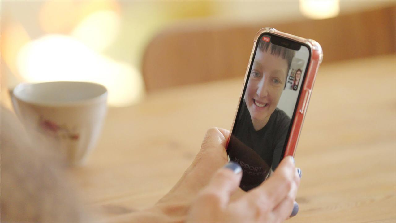 Das Bild zeigt eine Person, die ein Smartphone in der Hand hält. Auf dem Smartphone-Display ist eine Videoanruf-Situation zu sehen, in der die Person auf dem Bildschirm lächelt. Der Hintergrund ist unscharf, wobei helle, verschwommene Lichter erkennbar sind, die eine warme Atmosphäre schaffen. Auf dem Tisch vor der Person steht eine kleine, weiße Tasse, die mit einem sanften Blumenmotiv verziert ist. Die Hand, die das Telefon hält, hat gepflegte Nägel, und die Person scheint sich in einem gemütlichen, einladenden Raum zu befinden.