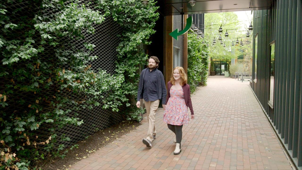 Das Bild zeigt ein Paar, das auf einem gepflasterten Weg geht, der von grünen Pflanzen und Büschen gesäumt ist. Der Weg scheint sich durch einen modernen, urbanen Bereich zu winden, wo sich auch Gebäude mit Fenstern auf der rechten Seite befinden. Über dem Weg hängen kleine Lichter, die eine einladende Atmosphäre erzeugen. Das Paar besteht aus einem Mann mit lockigem, dunklem Haar und einem Bart, der eine dunkelblaue Bluse und beige Hose trägt, und einer Frau mit langen, rotblonden Haaren, die ein blumiges Kleid und einen dunkel-lila Cardigan anhat. Beide wirken entspannt und lächeln, während sie sich unterhalten. Eine grüne Pfeil-Markierung an einer Wand zeigt in eine Richtung, und im Hintergrund sind weitere grüne Pflanzen zu sehen.