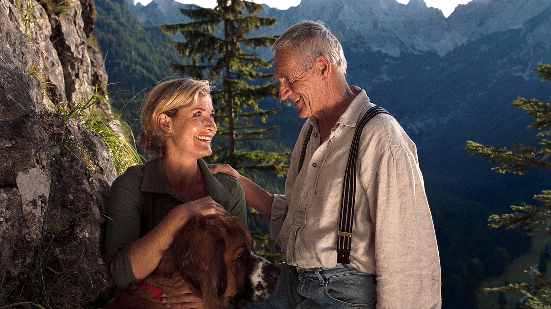 Eine Frau mit einem Bernhardiner im Arm und ein Mann vor einer Bergkulisse. 