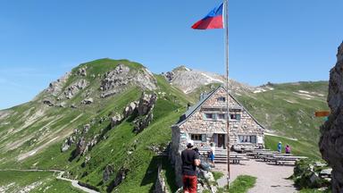 Das Bild zeigt eine alpine Landschaft in Liechtenstein mit der Pfälzerhütte, einem steinernen Gebäude, das sich auf einer Anhöhe befindet. Die Hütte hat ein geneigtes Dach und ist von mehreren Bänken und Tischen umgeben, die auf einer gepflasterten Fläche stehen. Im Hintergrund erheben sich grüne, bewaldete Berge, die teilweise mit Felsen und Schnee durchzogen sind. Am Gebäude weht eine Flagge, die in den Farben Blau und Rot gehalten ist. Der Himmel ist klar und sonnig. Einige Personen sind in der Nähe der Hütte zu sehen, die entweder herumlaufen oder die Aussicht genießen. Der Wanderweg, der sich durch die Landschaft schlängelt, ist in der Mitte des Bildes zu erkennen. Die Szene vermittelt eine ruhige, naturnahe Atmosphäre.