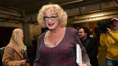 Ein als Frau gekleideter Travestiekünstler mit blonden Locken und dunkler Brille.