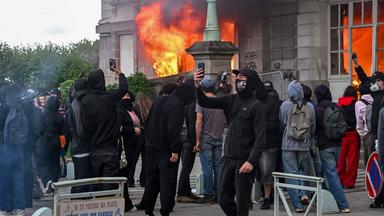 Schwarz gekleidete und vermummte Personen machen Selfies vor einem brennenden Gebäude.