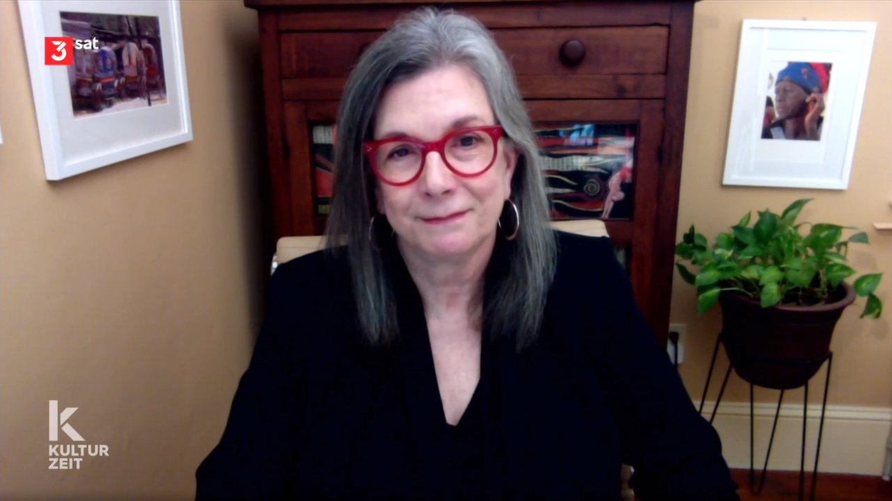 Eine Frau mit grauen Haaren und roter Brille sitzt in einem Zimmer mit beiger Wand vor einem braunen Schrank und einer Pflanze. 