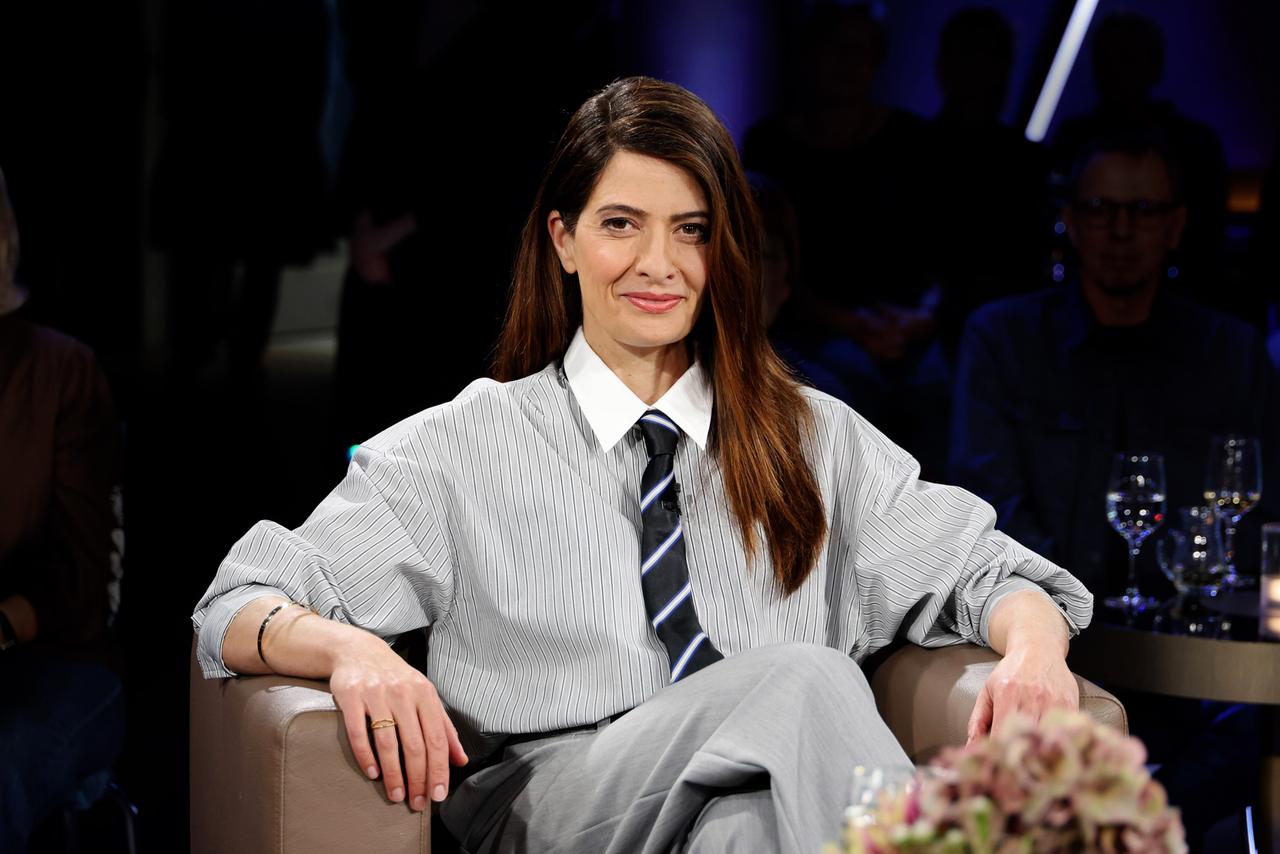 Eine Frau mit dunklen Haaren und freundlichem Gesichtsausdruck sitzt mit überschlagenen Beinen auf einem Sessel und blickt in Richtung des Betrachters.