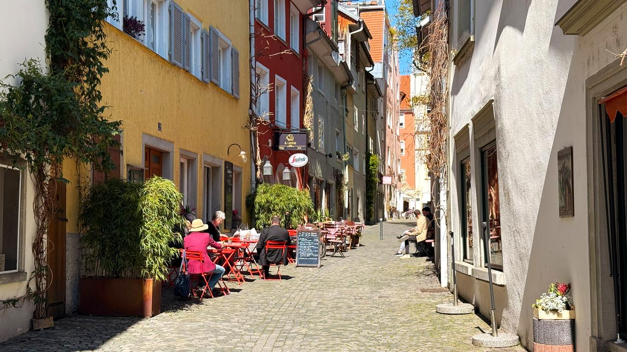 Das Bild zeigt eine enge Gasse mit bunten Gebäuden. An den Seiten stehen Tisch-Möbel in rot, an denen Personen sitzen. Es liegt gepflasterter Boden, und es sind Pflanzen sowie Dekorationen sichtbar.