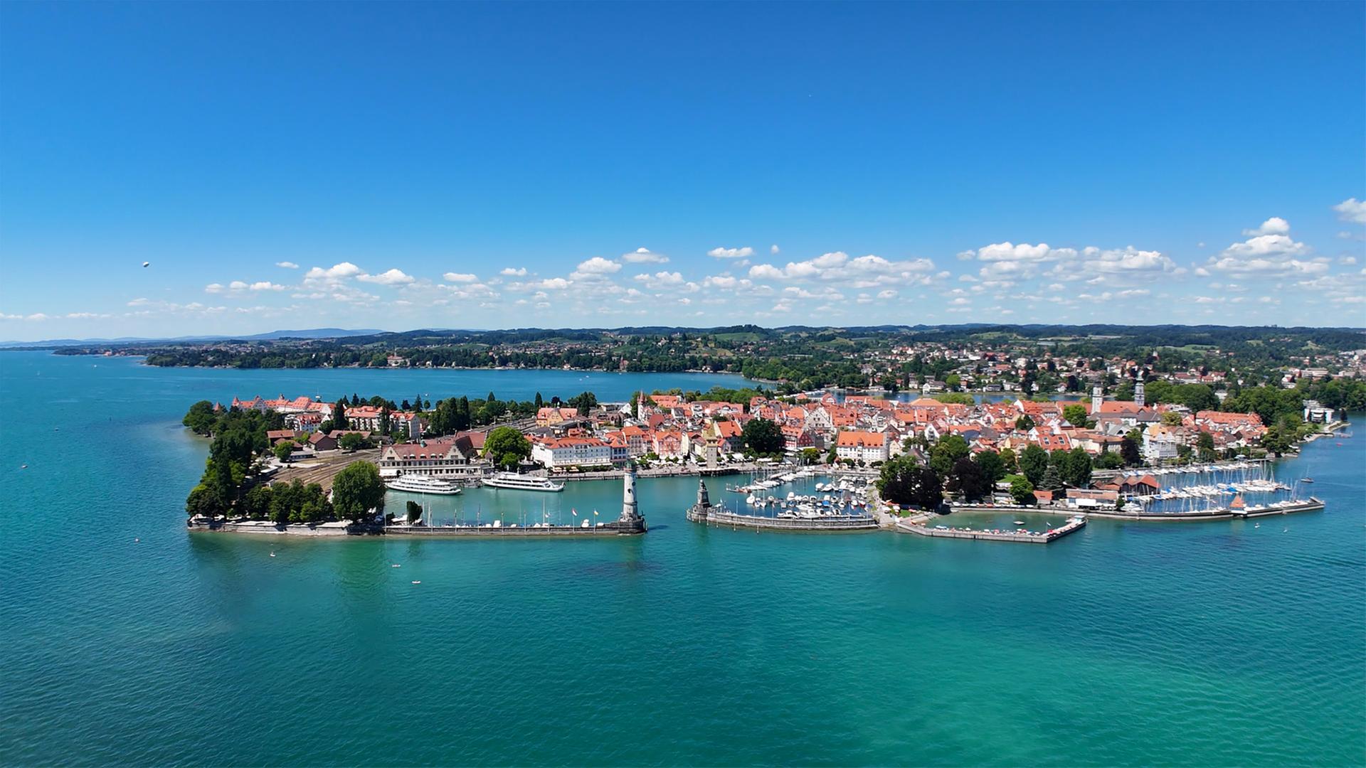 Das Bild zeigt die Insel Lindau im Bodensee. Im Vordergrund sind Wasserflächen und Häfen mit Booten zu sehen. Im Hintergrund erkennt man die Altstadt mit bunter Architektur und Bäumen. Der Himmel ist blau mit wenigen Wolken.