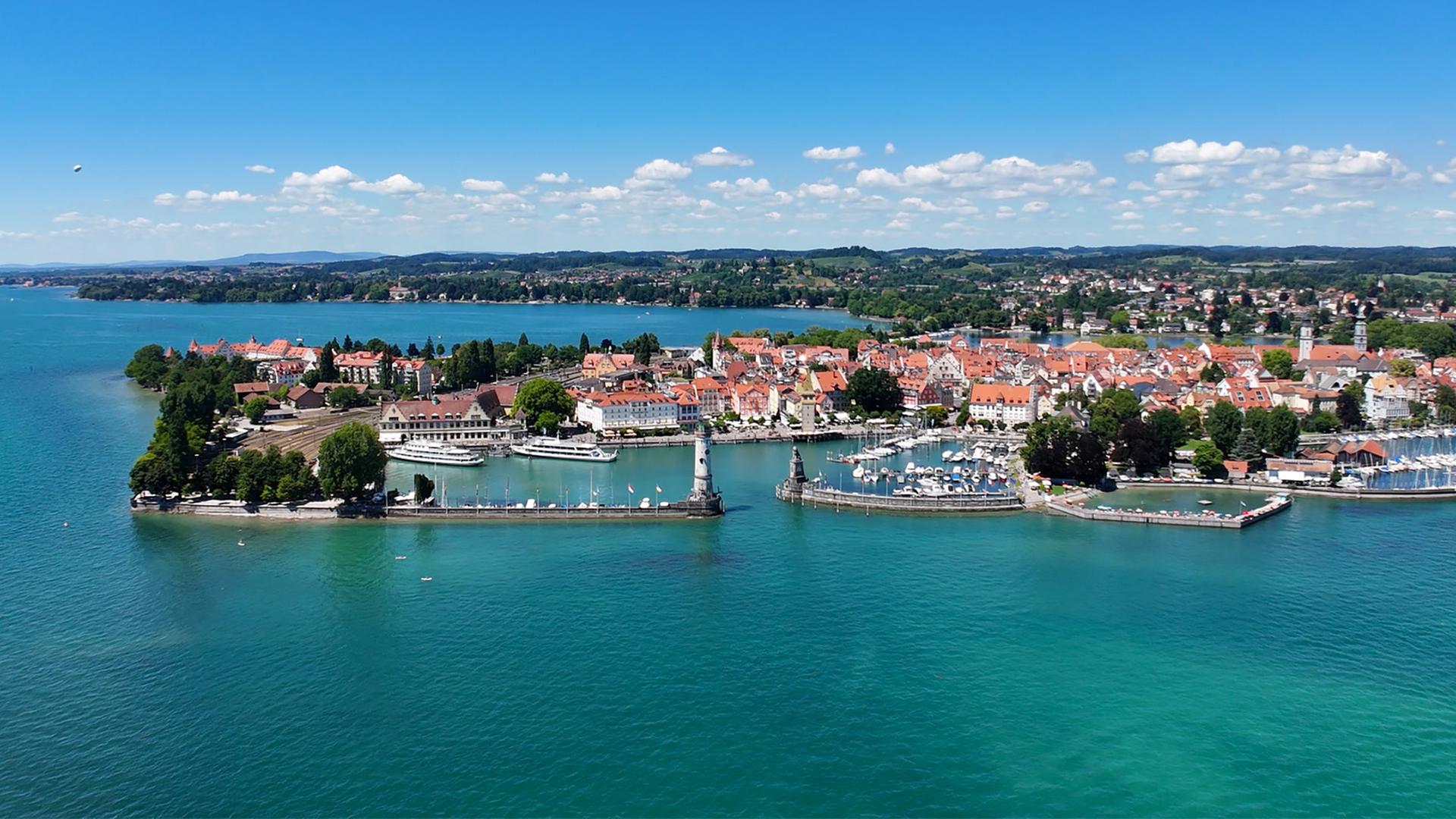 Das Bild zeigt die Insel Lindau im Bodensee. Im Vordergrund sind Wasserflächen und Häfen mit Booten zu sehen. Im Hintergrund erkennt man die Altstadt mit bunter Architektur und Bäumen. Der Himmel ist blau mit wenigen Wolken.