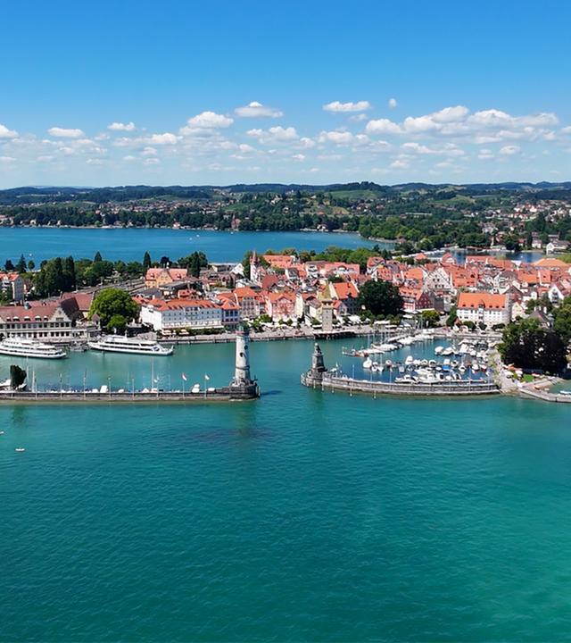 Das Bild zeigt die Insel Lindau im Bodensee. Im Vordergrund sind Wasserflächen und Häfen mit Booten zu sehen. Im Hintergrund erkennt man die Altstadt mit bunter Architektur und Bäumen. Der Himmel ist blau mit wenigen Wolken.
