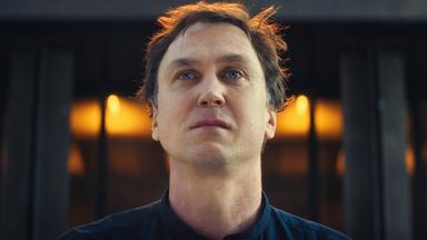 In einer Nahaufnahme blickt Tom Lunies (Lars Eidinger) mit feuchten Augen an der Kamera vorbei nach vorn. Die Umgebung liegt im Dunkeln. Hinter ihm leuchtet auf Augenhöhe ein goldener Lichtschein.