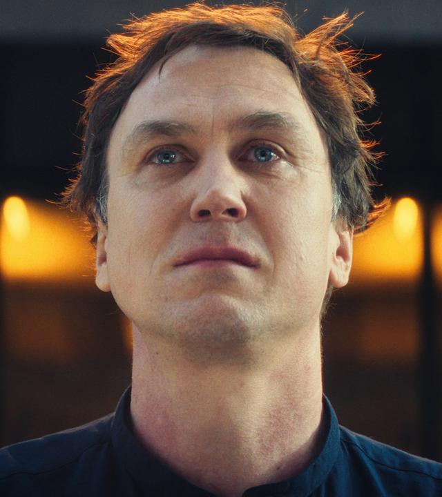 In einer Nahaufnahme blickt Tom Lunies (Lars Eidinger) mit feuchten Augen an der Kamera vorbei nach vorn. Die Umgebung liegt im Dunkeln. Hinter ihm leuchtet auf Augenhöhe ein goldener Lichtschein.