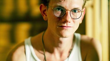 Ein junger Mann mit lockigem Haar und runden Brillen, in einem weißen Tanktop, sitzt in einem Raum mit verschwommenem Hintergrund.