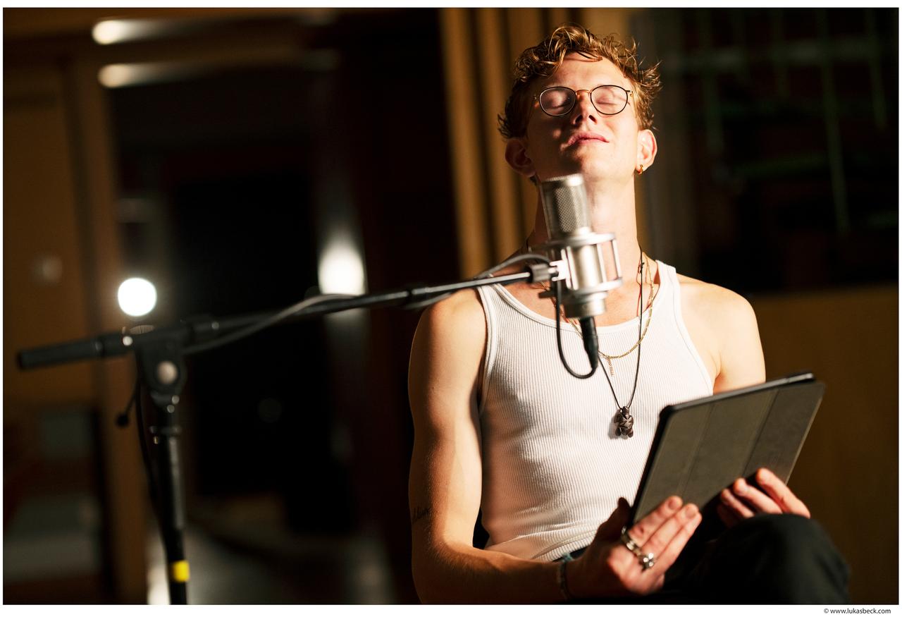 Ein junger Mann mit Brille sitzt vor einem Mikrofon in einem Tonstudio. Er trägt ein weißes Tanktop und hält ein Tablet in den Händen.