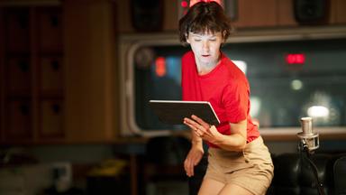 Eine Frau im roten T-Shirt und beige Shorts sitzt in einem Raum mit Studioequipment und liest von einem Tablet.