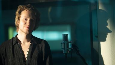 Nils Arztmann steht in einem Tonstudio vor einem Mikrofon, im Hintergrund ist sein Schatten an der Wand sichtbar.