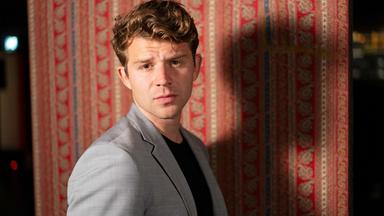 Ein Mann im hellgrauen Sakko steht vor einem bunten Teppich und blickt ernst in die Kamera.