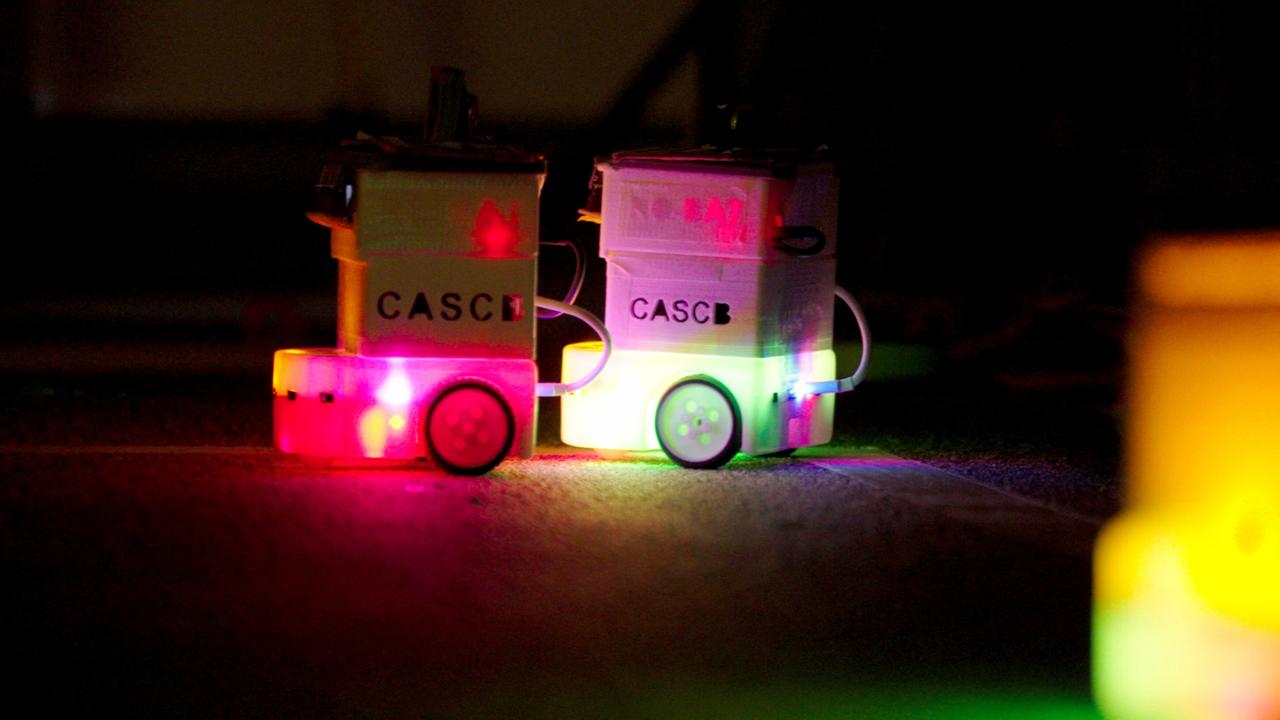 Das Bild zeigt zwei kleine Roboter, die im Vordergrund stehen. Sie haben eine kompakte, kastenförmige Bauweise und sind mit bunten Lichtern ausgestattet, die in verschiedenen Farben leuchten, darunter Rot, Blau, Gelb und Grün. Auf der Vorderseite der Roboter ist der Schriftzug „CASC“ zu sehen. Die Roboter bewegen sich auf einer dunklen Oberfläche, was die Helligkeit der Lichter verstärkt. Der Hintergrund ist unscharf und dunkel, was den Fokus auf die Roboter lenkt. Die Szene vermittelt einen Eindruck von Technologie und Innovation, möglicherweise im Kontext eines Forschungsprojekts.