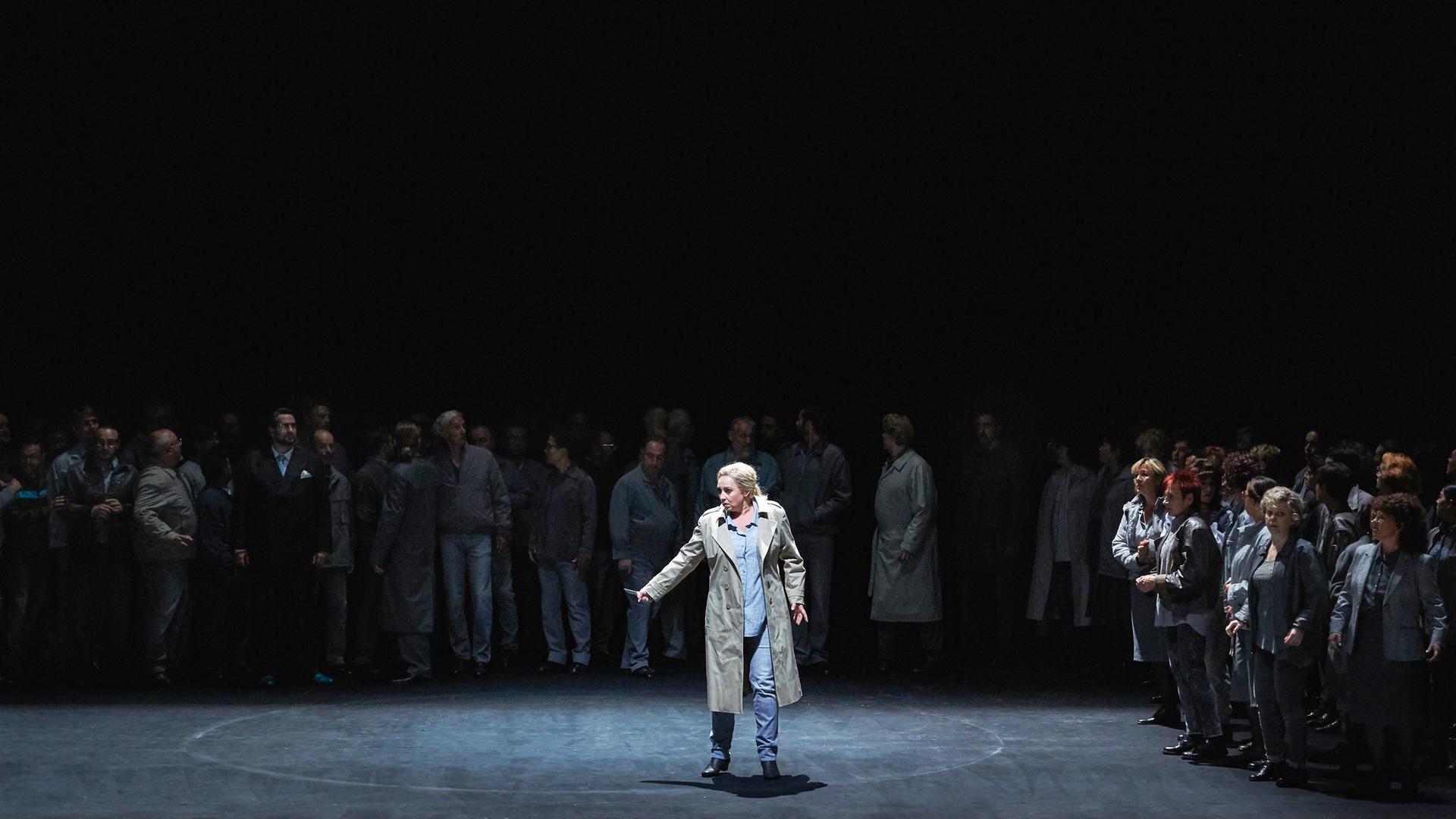 Szene auf einer Opernbühne. Im Vordergrund steht eine Frau im Trenchcoat im Licht, dahinter viele Menschen im Dunkeln.