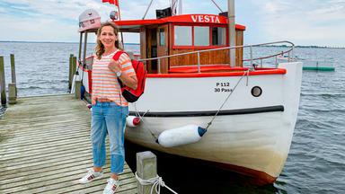 Die Moderatorin steht lächelnd mit einem roten Rucksack vor einem kleinen Kutter.