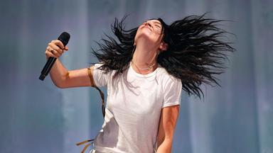 Eine Frau mit dunklen langen Haaren Hält ein Mikrofon in der Hand und schüttelt ihre lange Mähne.