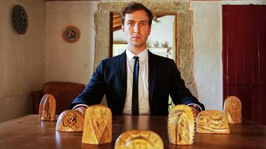 Ein Mann im Anzug sitzt an einem Tisch, auf dem Holzfiguren stehen